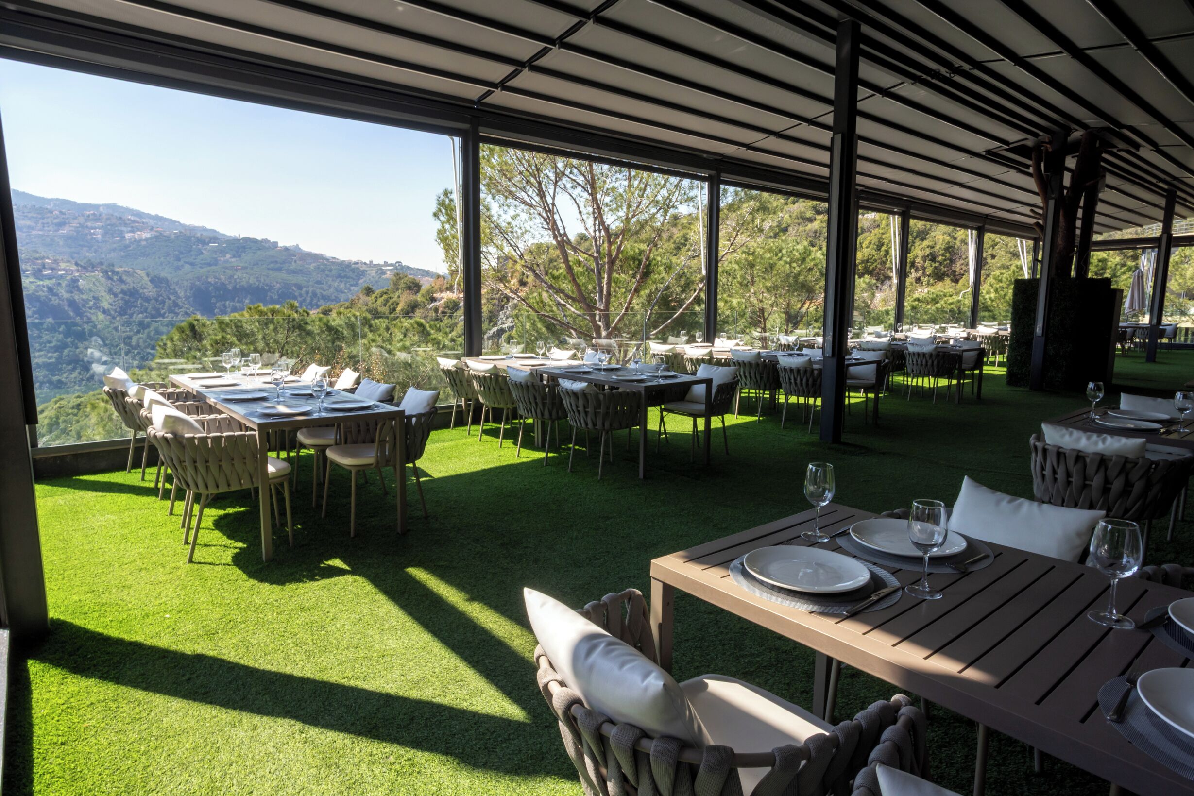 Un área de comedor al aire libre con mesas y sillas, rodeada por una estructura cubierta con ventanas grandes que ofrecen vistas a las colinas.