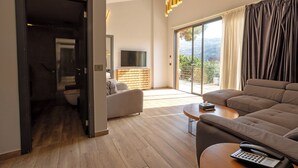 A modern living room with a sofa, a coffee table, and a TV. There is a sliding glass door leading to a balcony with a view of the outdoors. - Scappa Resort (Ajaltoun)