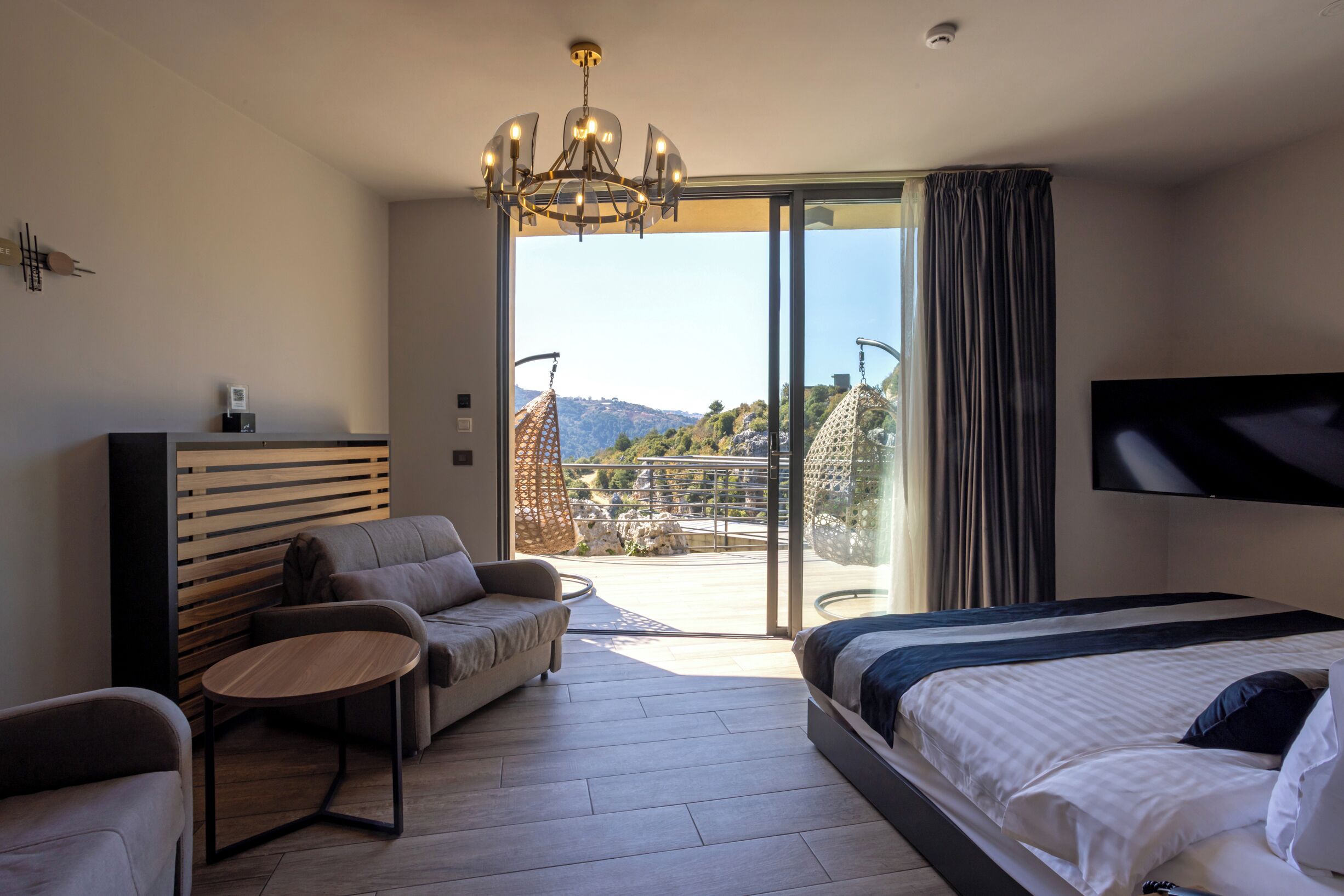 A modern hotel room with a large bed, a sofa, a small table, and a TV. The room has a balcony with a view of the mountains.