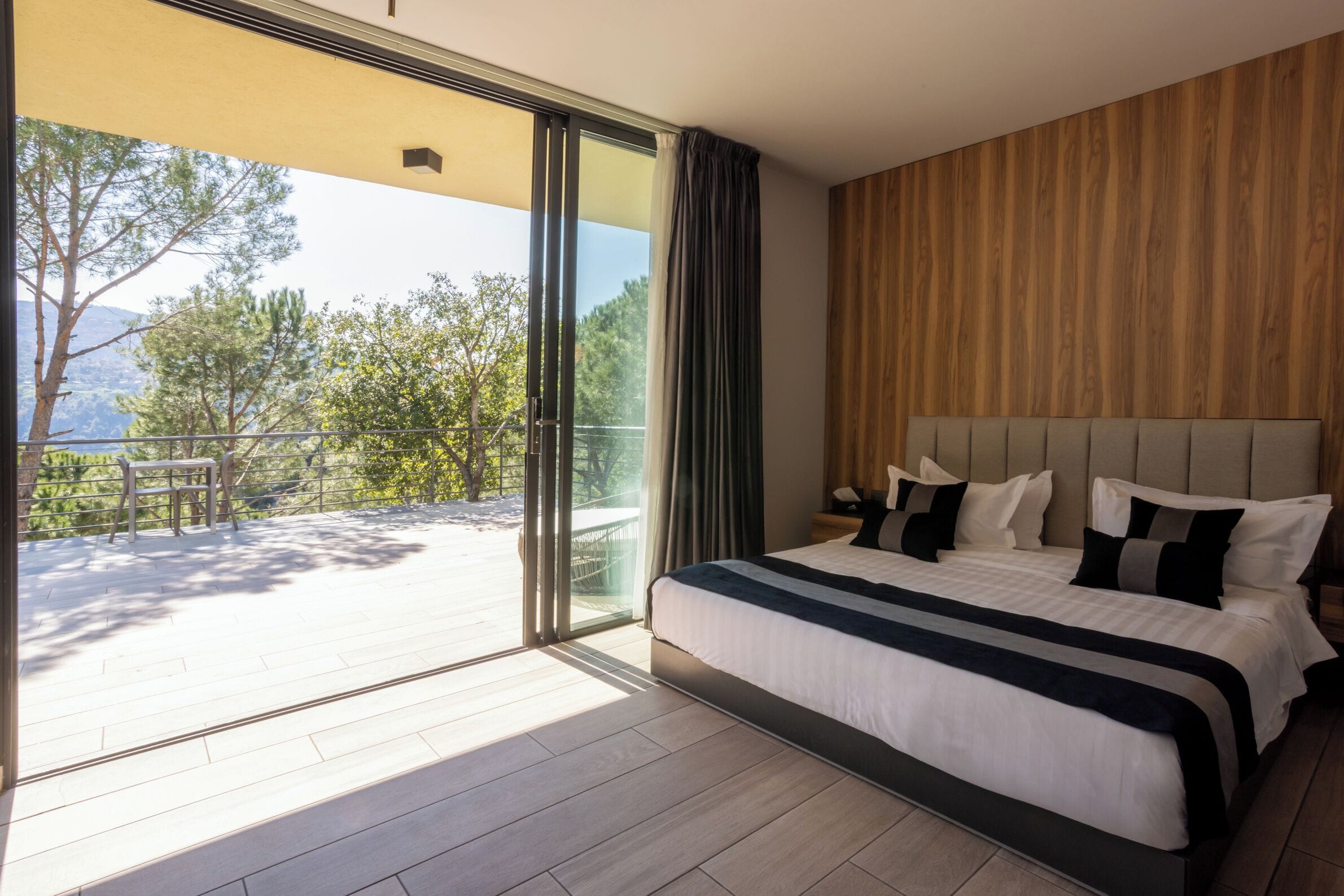 Un dormitorio moderno con una cama grande, cabecera de madera y un balcón con vistas a árboles y montañas.