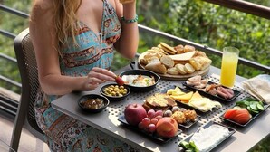 A person sitting at an outdoor table with a variety of food items and drinks, overlooking a mountainous landscape. - Scappa Resort (Ajaltoun)