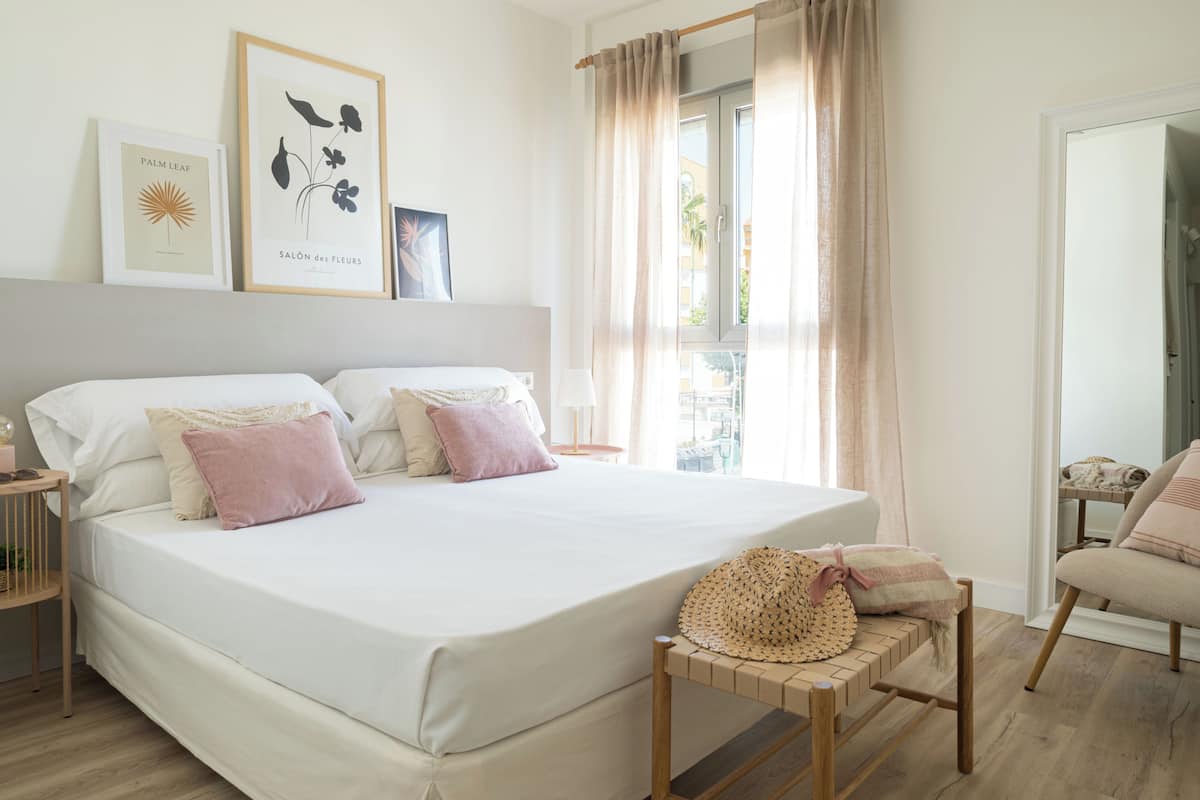 Un lit bien fait, agrémenté d’un arrangement d’oreillers blancs et roses, une table de chevet avec une lampe, et une estampe botanique encadrée au mur.