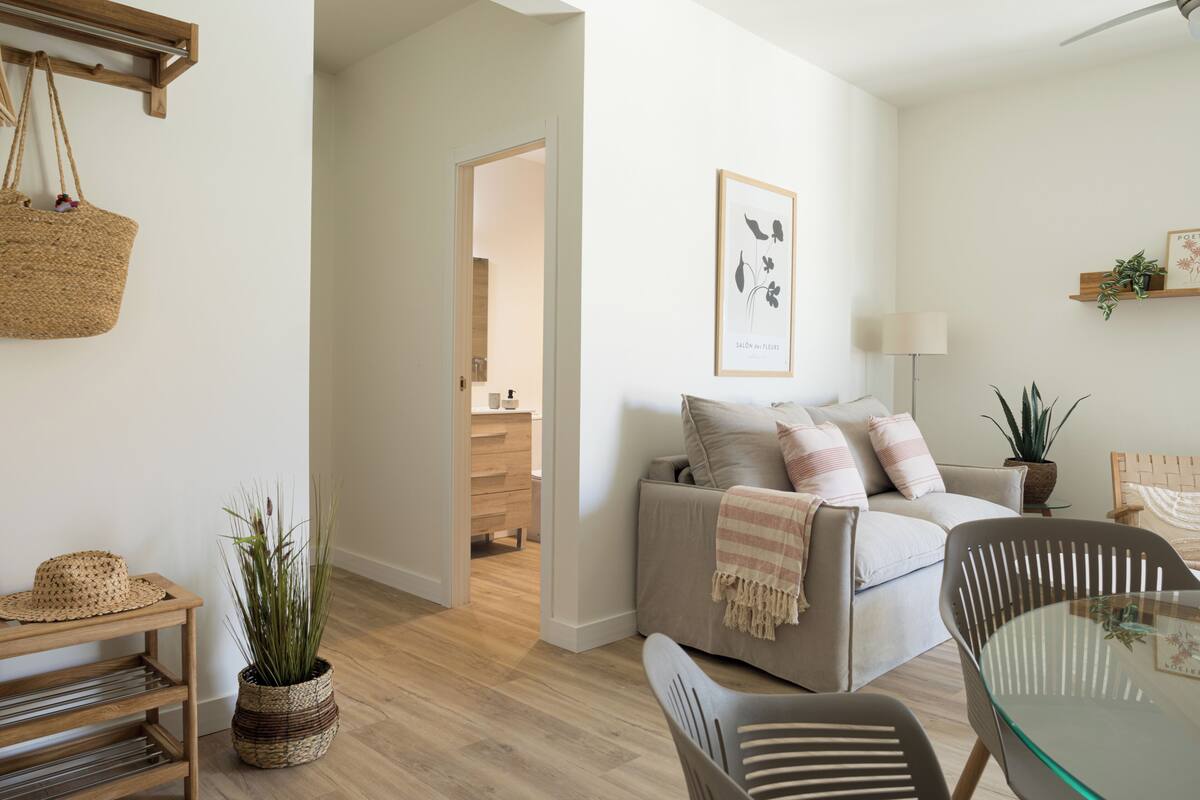 Un salon moderne avec un canapé, une table basse en verre et un coin repas avec des chaises. On y trouve une étagère en bois ornée d’une plante et un tableau encadré accroché au mur.