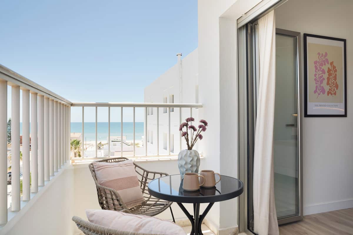 Un balcon avec vue sur la plage, des meubles en osier, une table avec deux tasses et une œuvre d’art encadrée accrochée au mur.