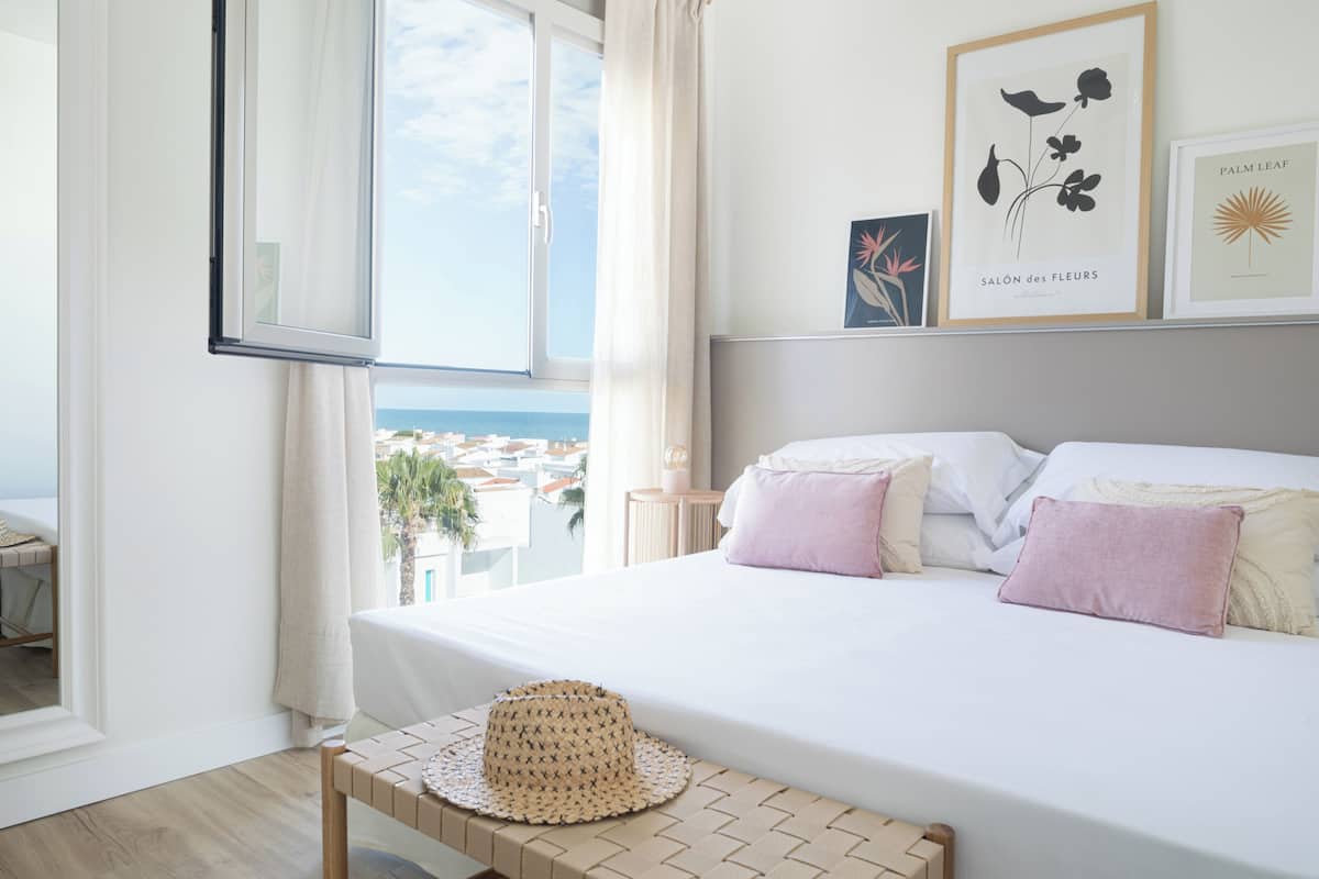 Une chambre avec un grand lit, une vue sur la mer et des œuvres d’art encadrées accrochées au mur.