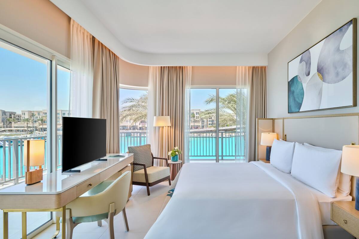 A hotel room with a large bed, a desk with a TV, a chair, and a view of a body of water.