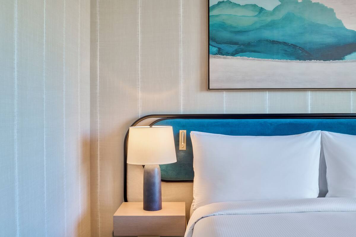 A neatly made bed with white linens, a bedside lamp, and a framed abstract painting on the wall.