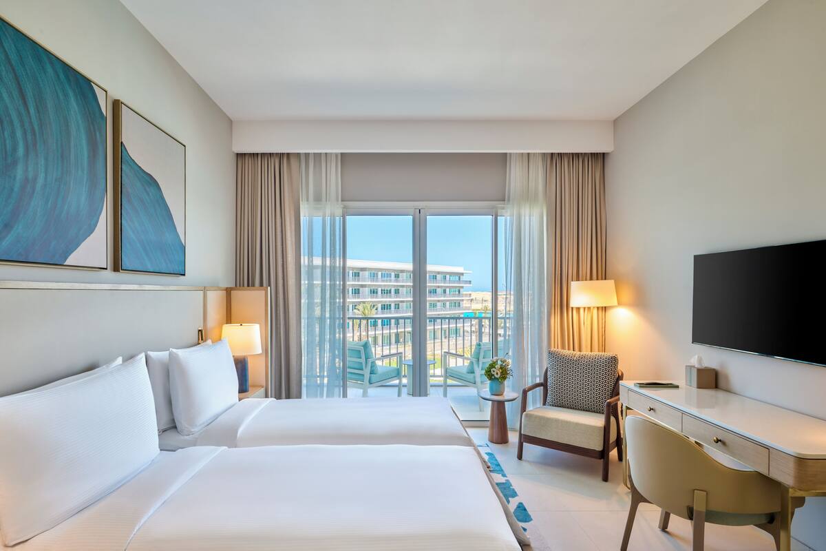 A hotel room with a large bed, a desk, a chair, a TV, and a balcony with a view of buildings.