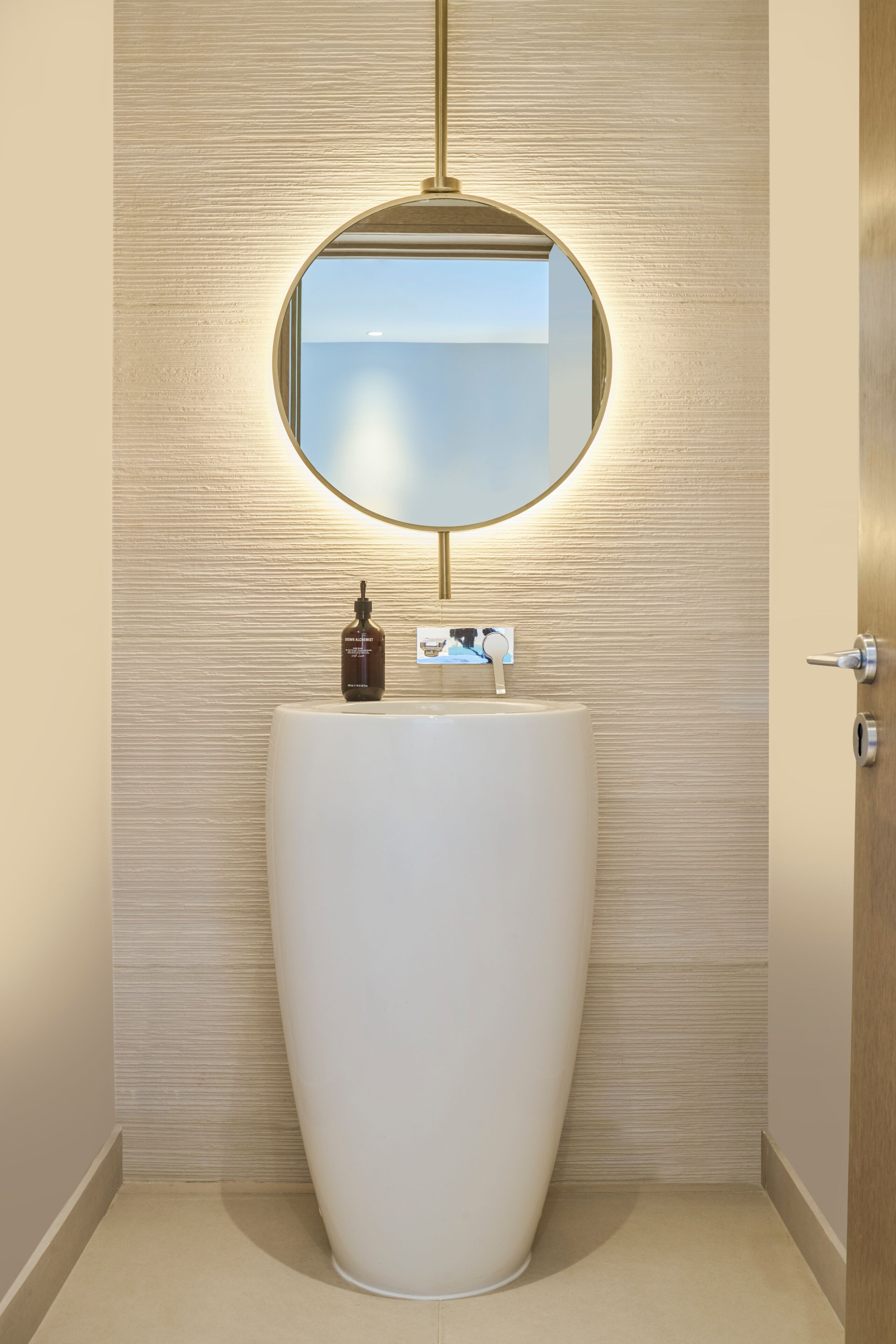 A modern bathroom with a large, white, oval-shaped sink, a round mirror with a gold frame, and a bottle of soap on the sink.