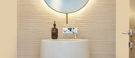 A modern bathroom with a large, white, oval-shaped sink, a round mirror with a gold frame, and a bottle of soap on the sink.