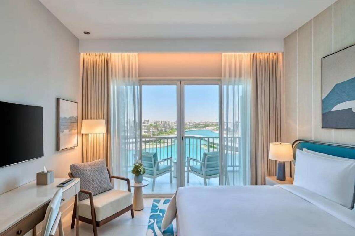 A hotel room with a bed, a desk, a chair, a TV, a balcony with a pool, and a city view.