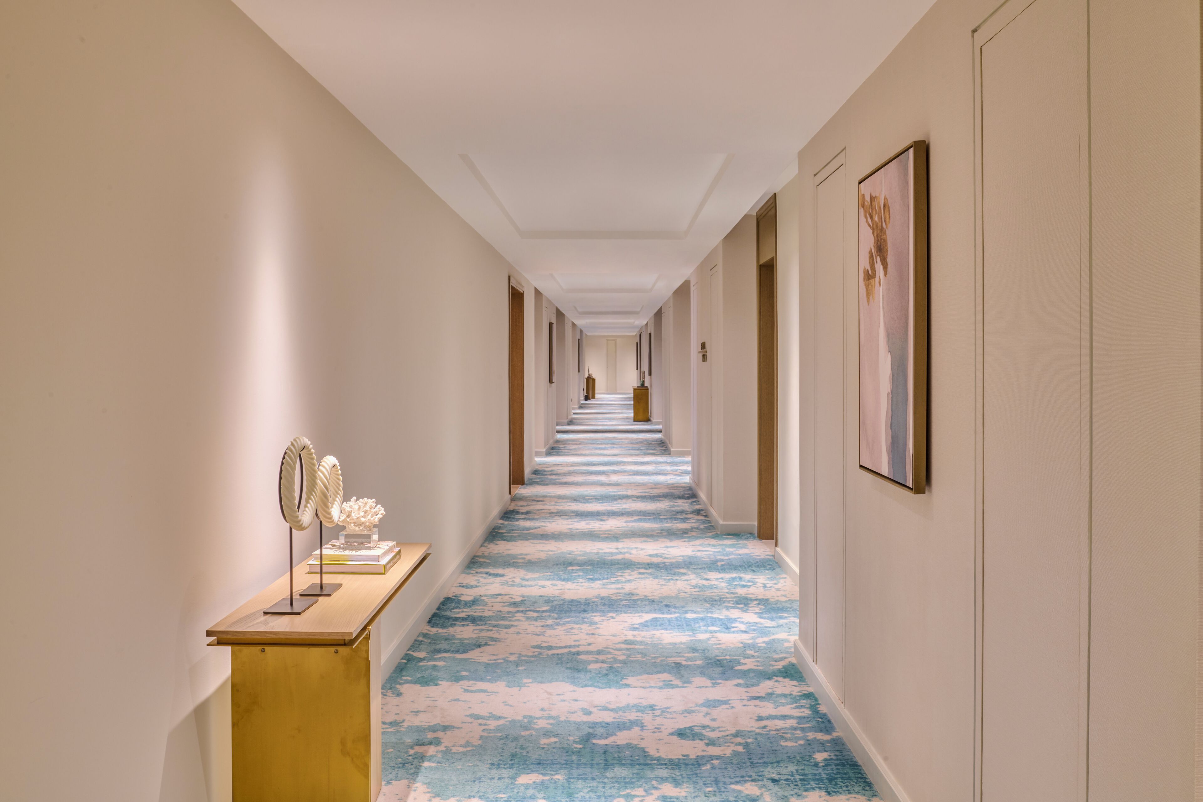 A hotel hallway with a blue carpet, framed artwork on the walls, and a reception desk with decorative items.