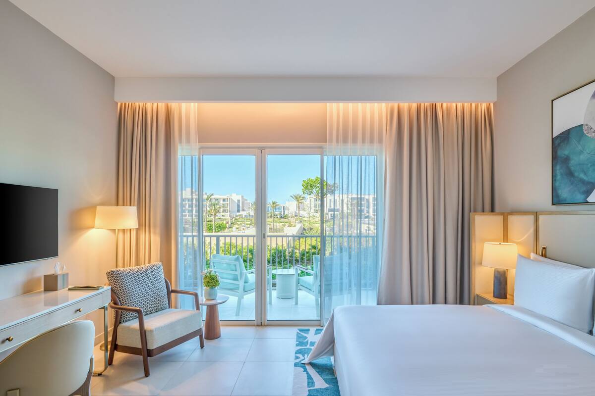 A hotel room with a bed, a desk, a chair, a TV, a balcony with a view of the city, and a painting on the wall.