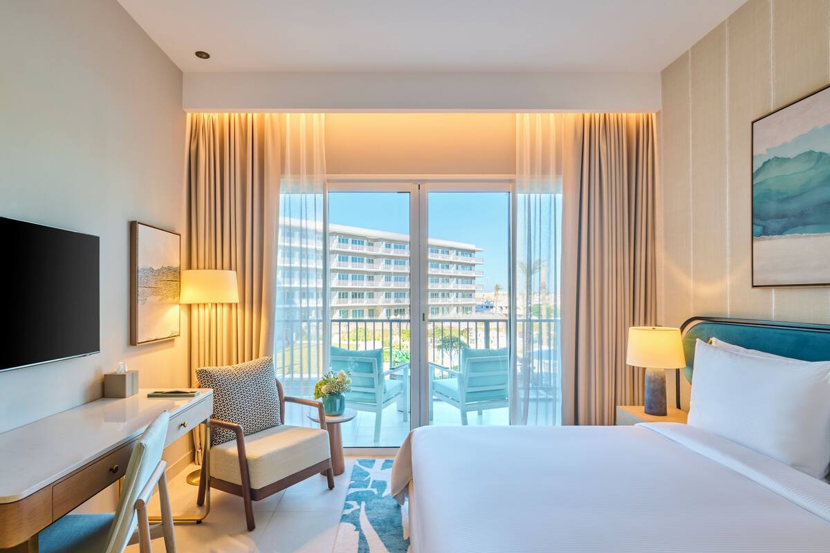A hotel room with a bed, a desk, a chair, a TV, and a balcony with a view of a building.