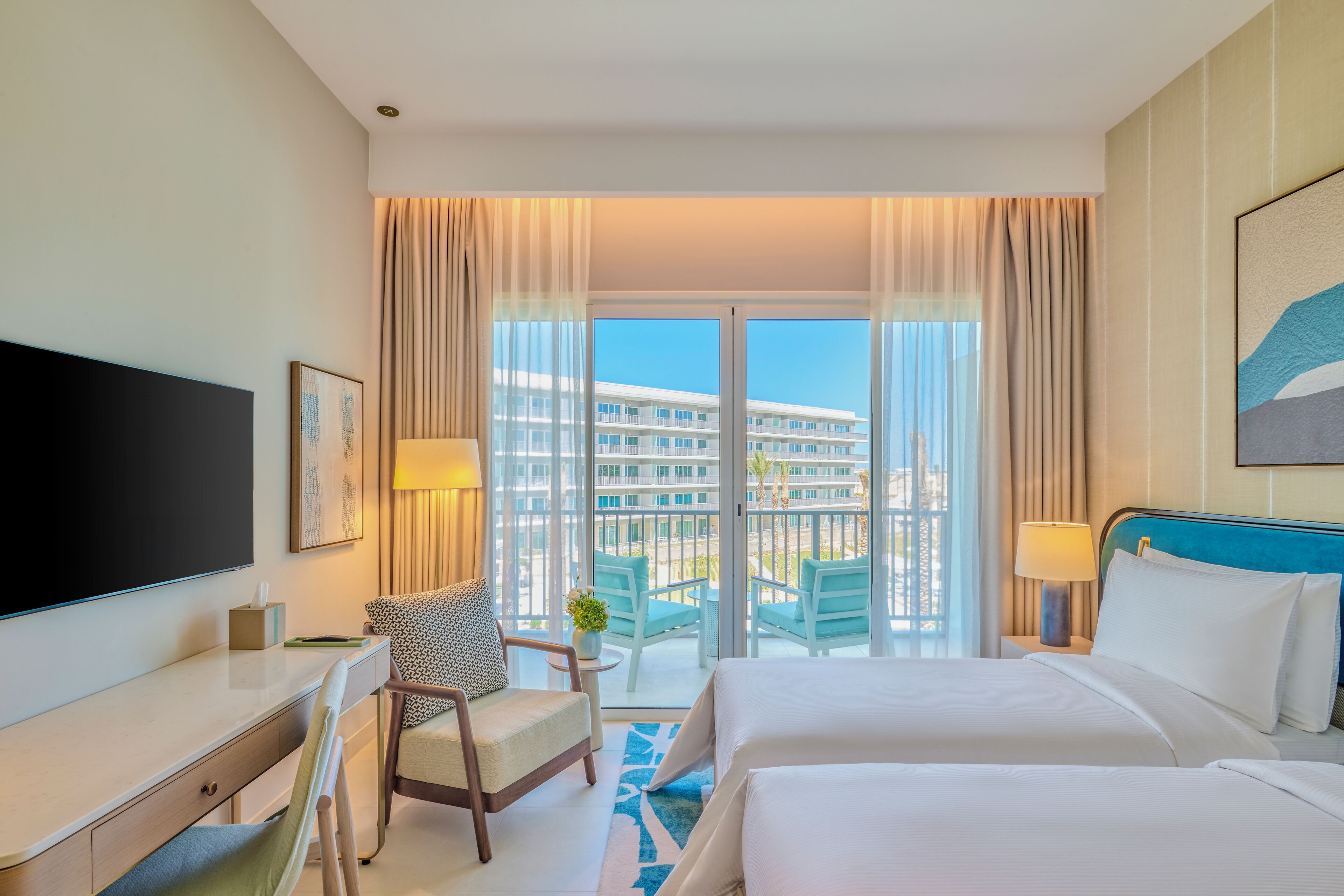 A hotel room with a large bed, a desk, a chair, a TV, and a balcony with a view of a building.