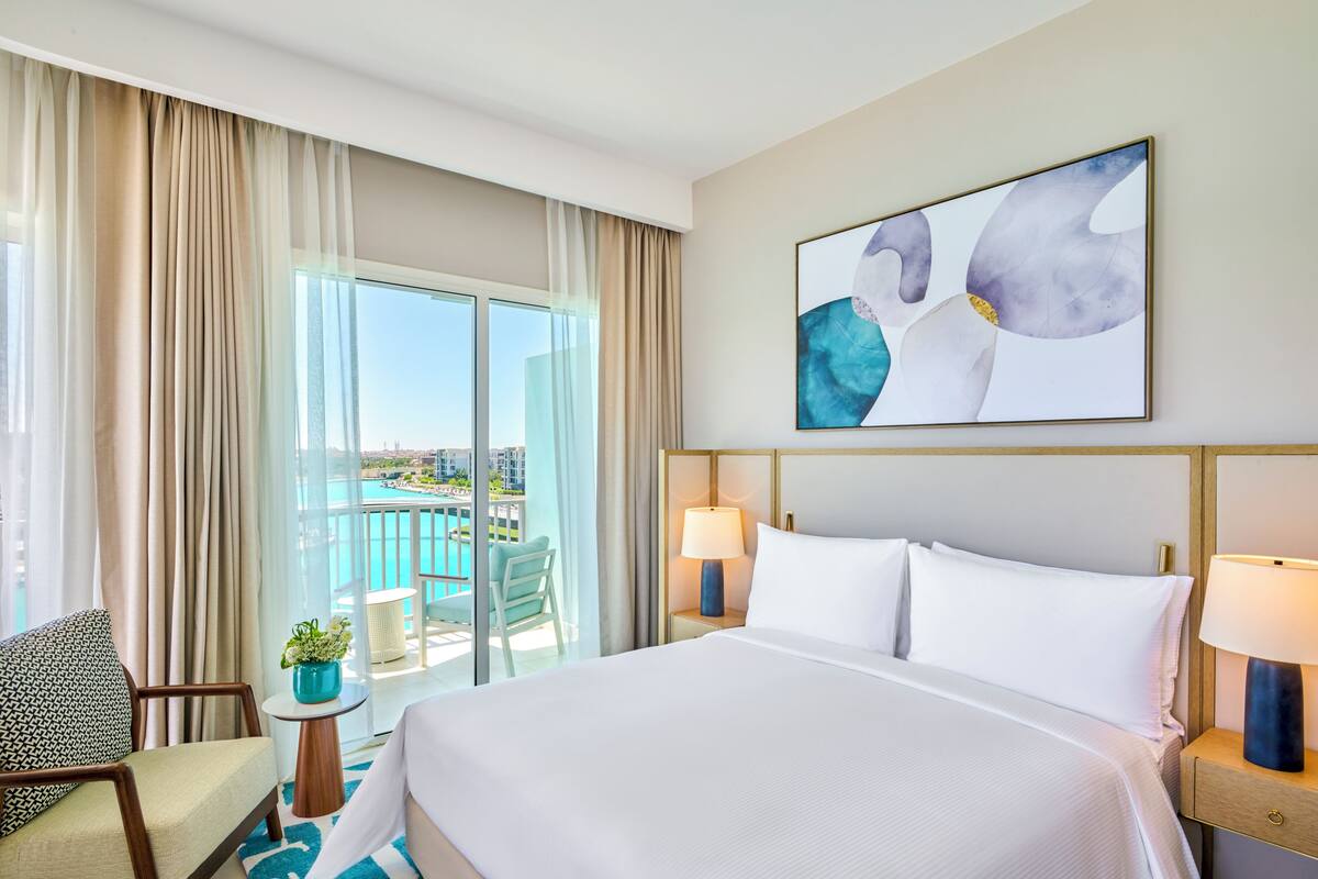 A hotel room with a large bed, two bedside lamps, a view of a pool and cityscape, and a painting on the wall.