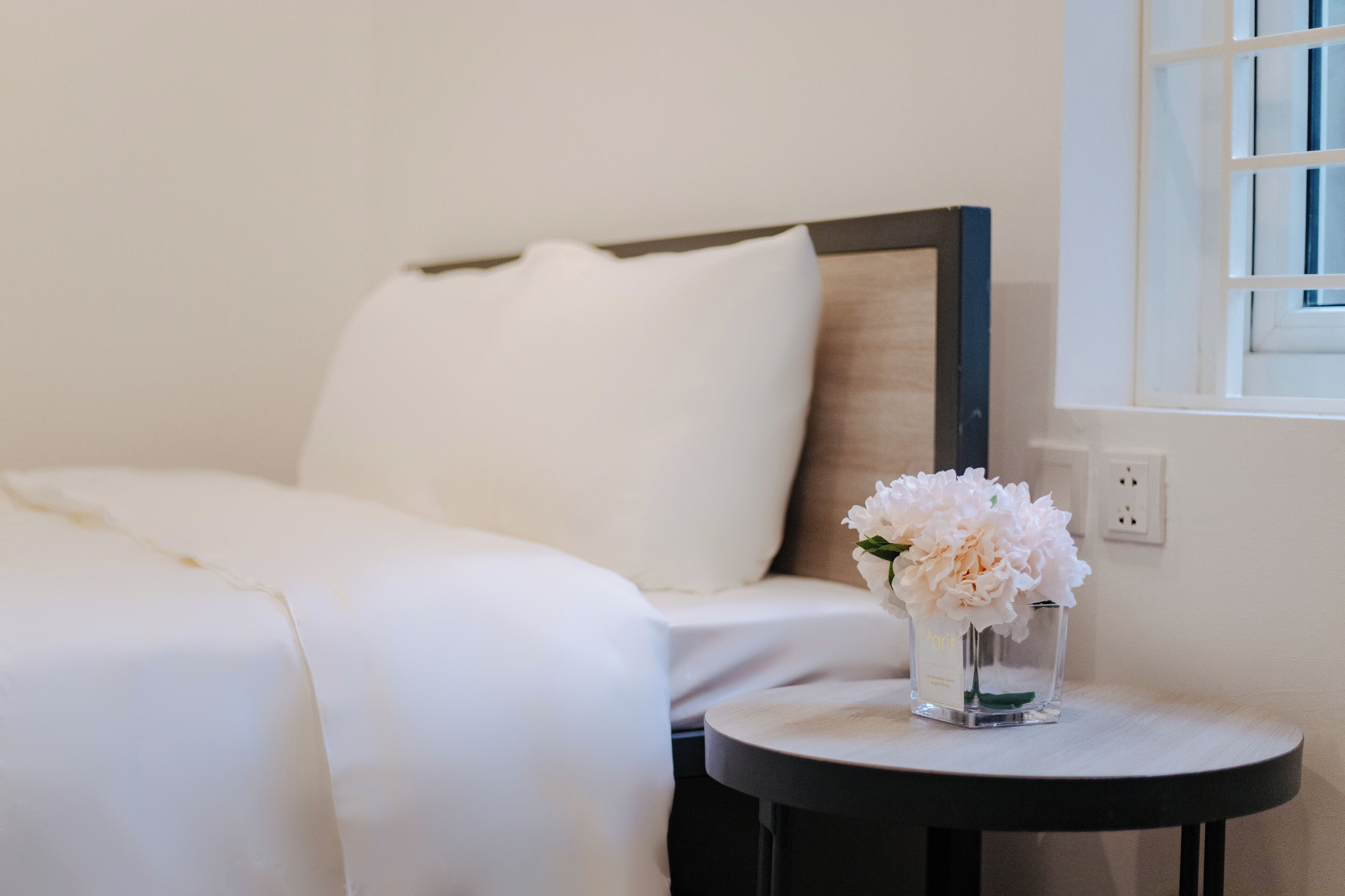 A neatly made bed with white bedding, a small round table with a vase of white flowers, and a window with a white frame.
