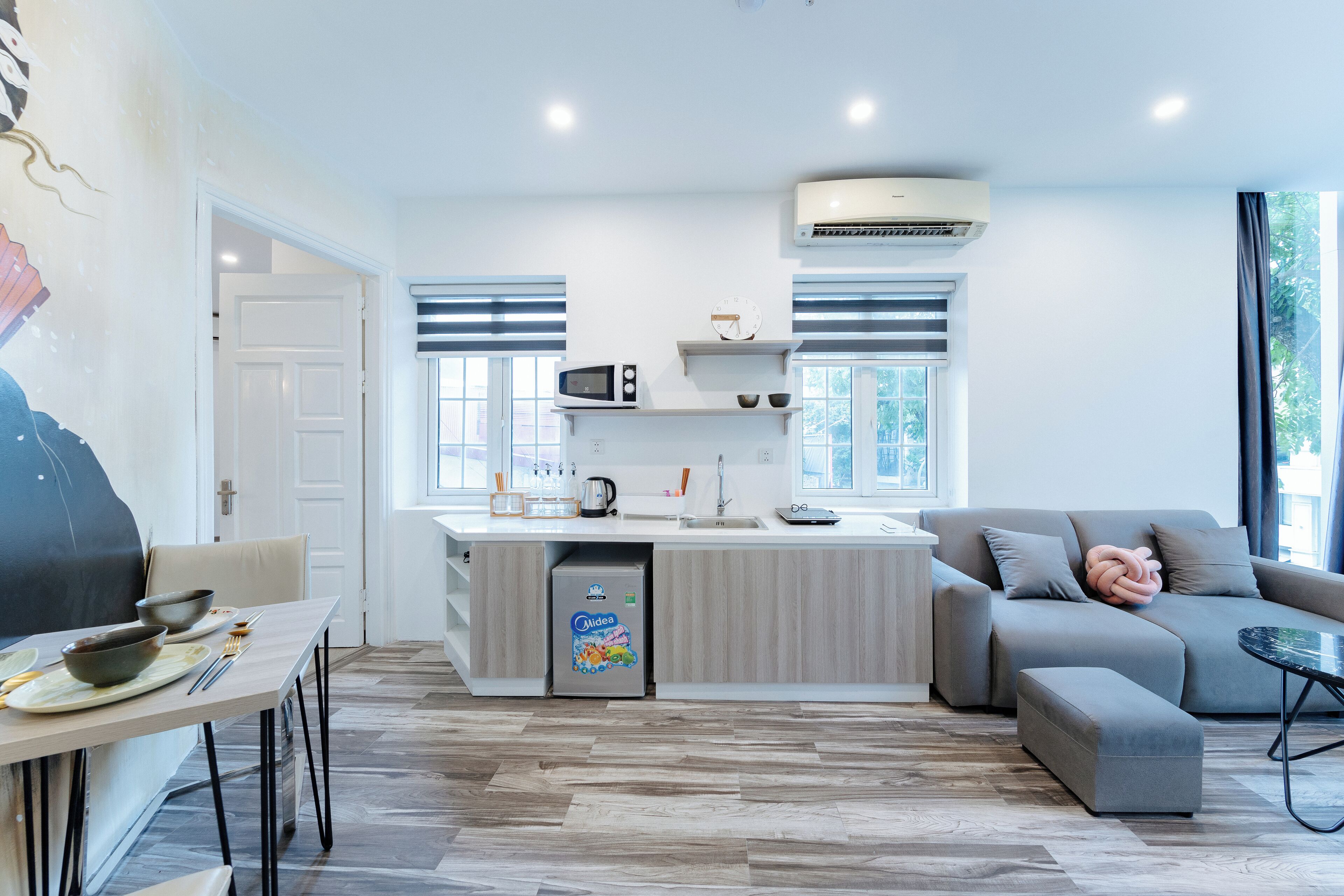Una cocina moderna con microondas integrado, refrigerador y lavamanos. Hay un sofá gris y una pequeña mesa con un objeto decorativo.