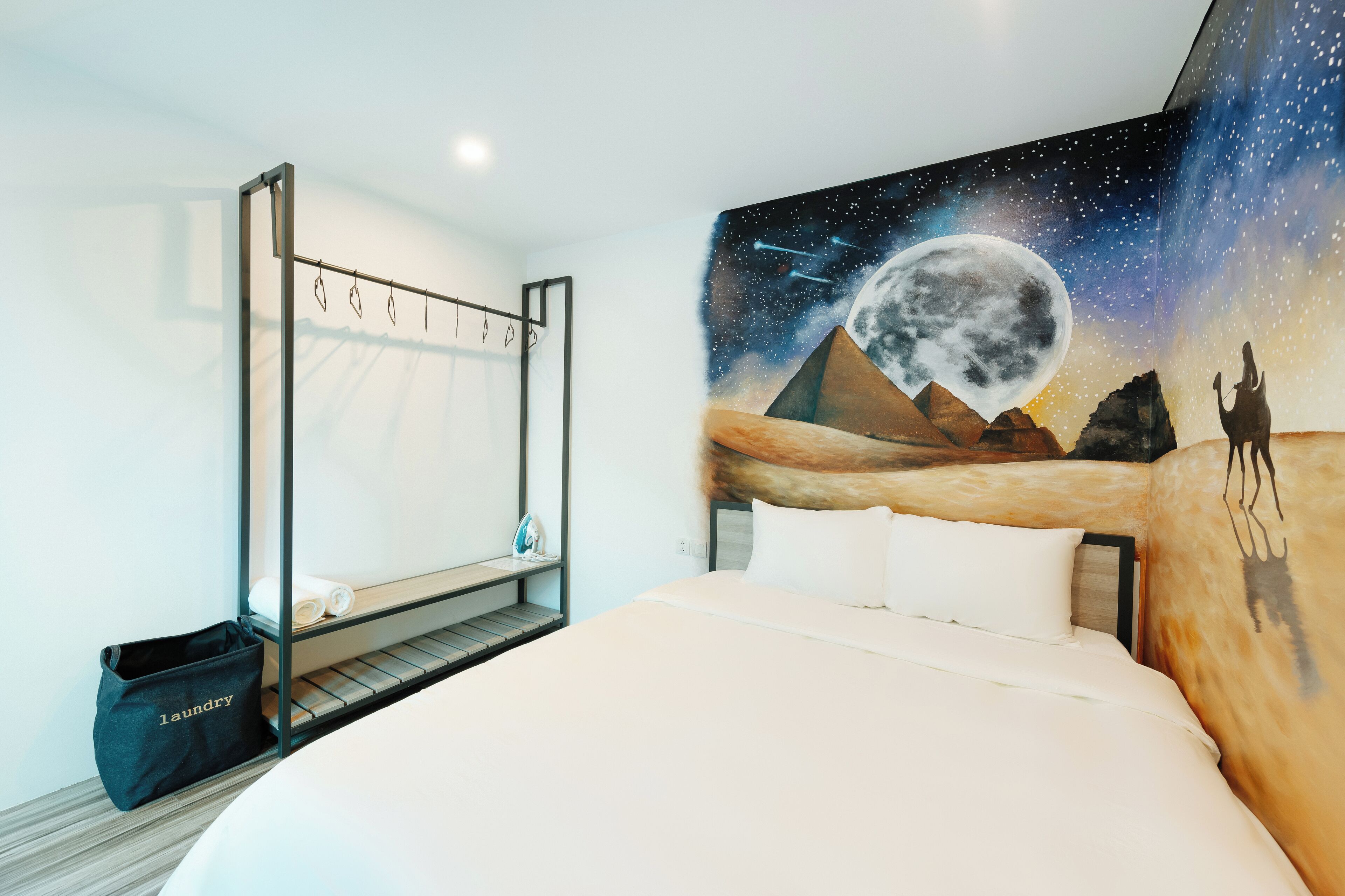 A bedroom with a bed, a hanging laundry rack, and a mural of a desert landscape with a moon.