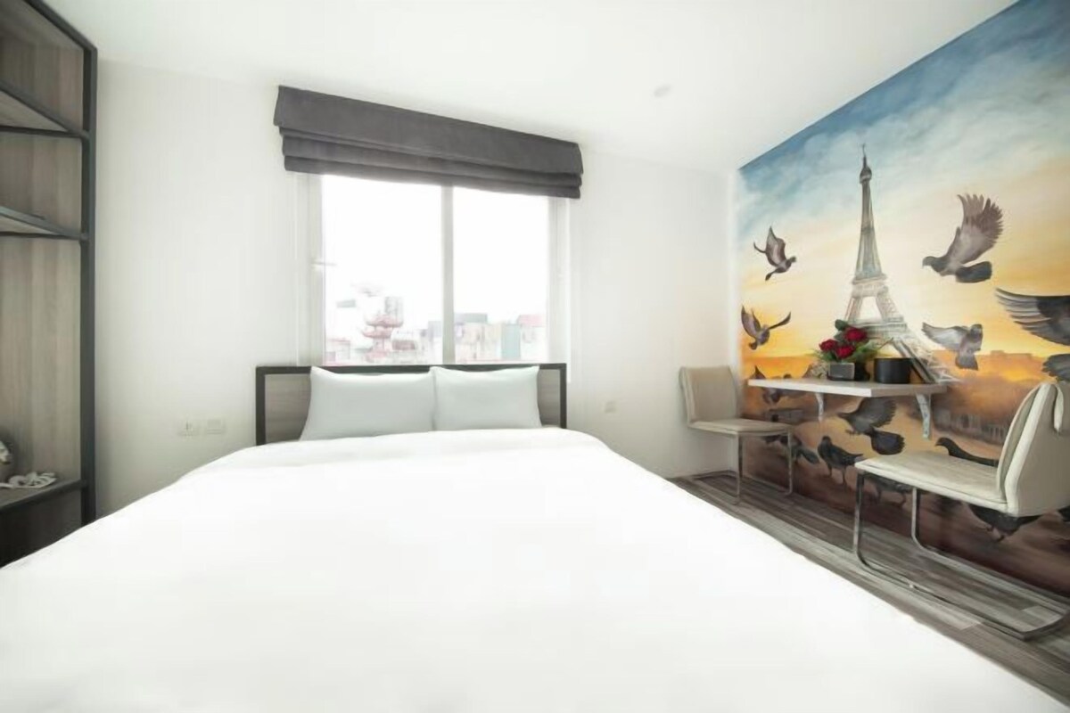 A hotel room with a large bed, a window with a view of the city, a painting of the Eiffel Tower, and a small table with chairs.