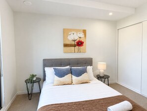 A hotel room with a bed, a bedside lamp, a small table with a plant, and a painting on the wall.