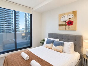 A hotel room with a large window, a bed with white bedding, a grey headboard, and a painting of flowers on the wall.