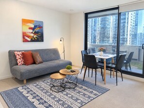 A modern living room with a grey sofa, a small round table with a plant, a dining area with a white table and black chairs, and a large window with a city view.