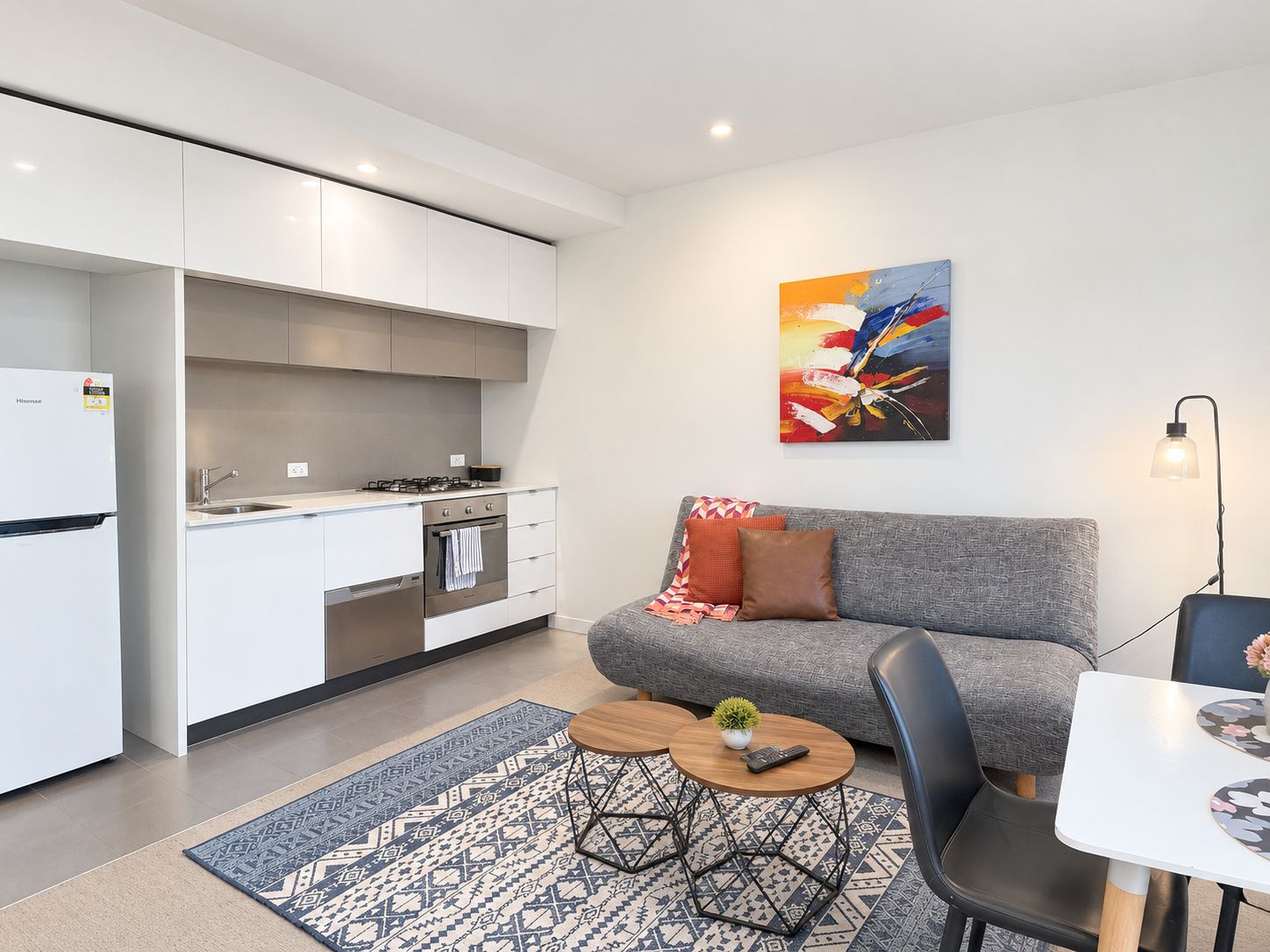 Eine moderne Küche mit weißen Schränken, einem Herd und einem Spülbecken. Es gibt ein graues Sofa mit einem farbenfrohen Kissen, einen kleinen runden Tisch mit einer Pflanze und einen schwarzen Stuhl. Ein Gemälde hängt an der Wand über dem Sofa.