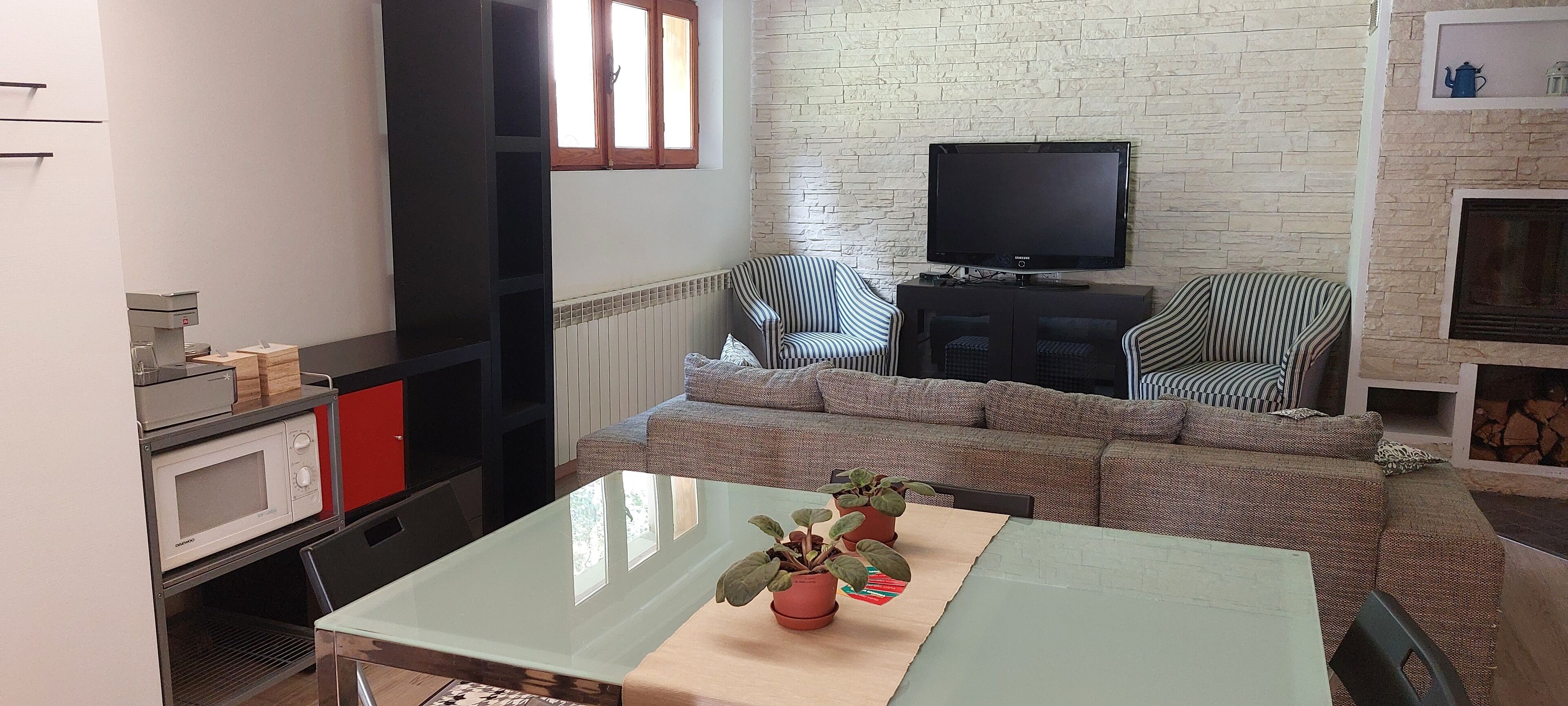 Un espace de vie compact comprenant un ensemble canapé, un téléviseur à écran plat fixé au mur, une table à manger avec des chaises et un coin cuisine équipé d’un four à micro-ondes et d’une machine à café.