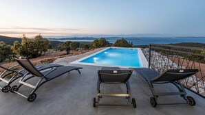 A rectangular pool with three lounge chairs on a concrete deck, overlooking a scenic view of the sea and hills.