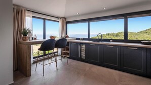 A modern kitchen with large windows, a view of the sea, and two black bar stools.