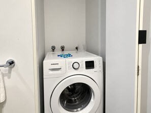 Une machine à laver Samsung dans une pièce blanche, avec une poignée de porte et une serviette accrochée à la porte.