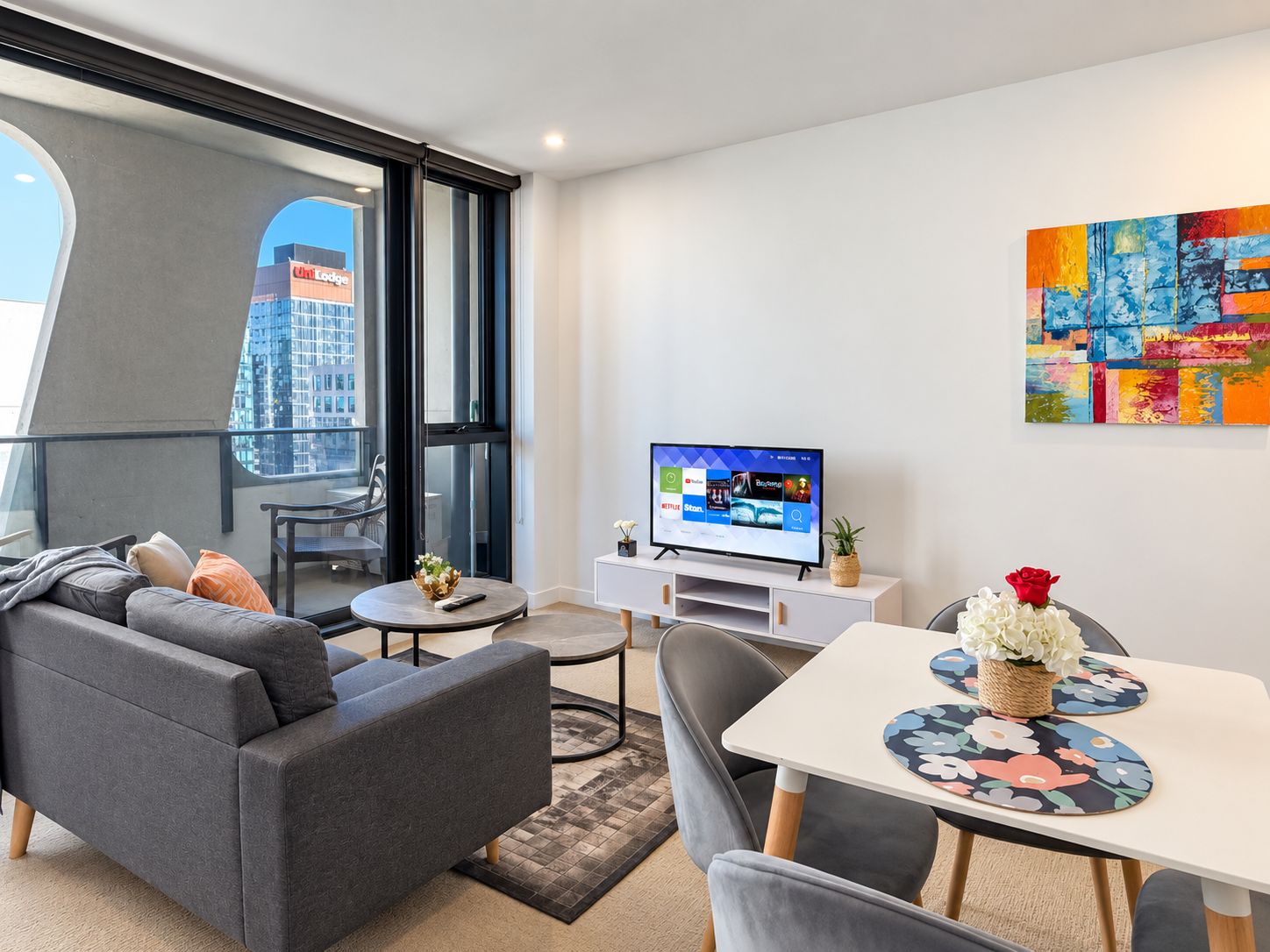 Una sala de estar moderna con un sofá gris, una mesa de comedor con individuales florales, un televisor de pantalla plana y un cuadro abstracto colorido en la pared.