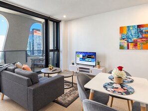 A modern living room with a grey sofa, a dining table with floral placemats, a flat-screen TV, and a colorful abstract painting on the wall.