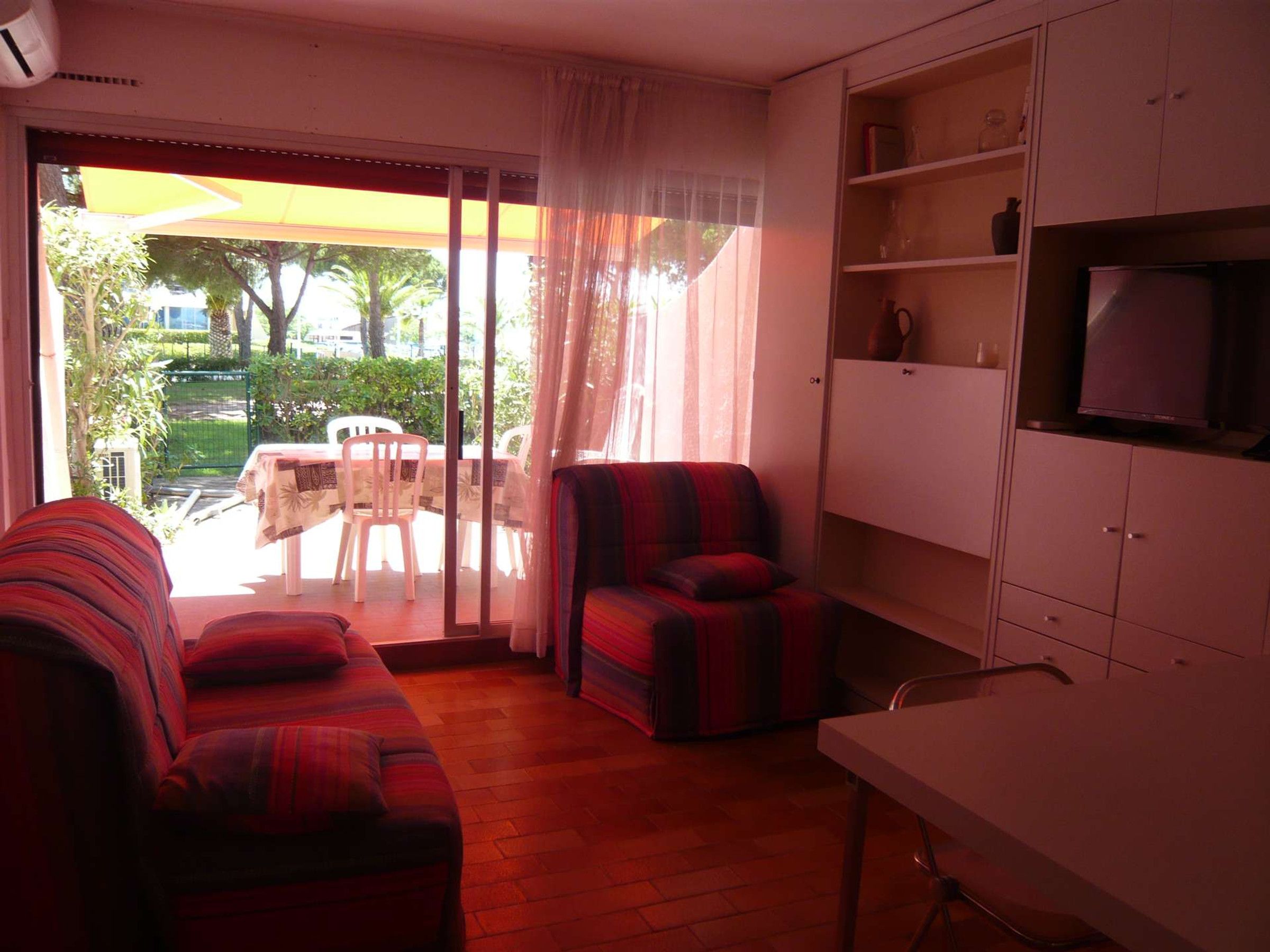 A living room with a sofa, armchair, and a TV on a cabinet. There is a sliding glass door leading to an outdoor area with a table and chairs.