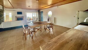 Een ruime kamer met houten plafond, een eettafel met stoelen, een keukenhoek en een uitzicht door een glazen deur op een buitenbalkon.