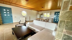 Een woonkamer met een bank, salontafel, stoel en een eettafel. Op de achtergrond een keuken met een fornuis, spoelbak en kasten.
