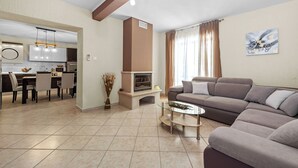 A modern living room with a sofa, a glass coffee table, and a fireplace. There is a dining area with chairs and a table, and a kitchen in the background. - Villa Ancora With Private Pool (Marcana)