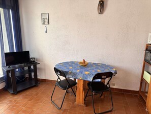 A small dining area with a round table covered by a blue tablecloth with a fruit pattern, two black chairs, a television on a stand, and a shelf with a basket. - Cozy Studio with Pool, Terrace & Private Parking in Le Grau-du-Roi (Port Camargue)