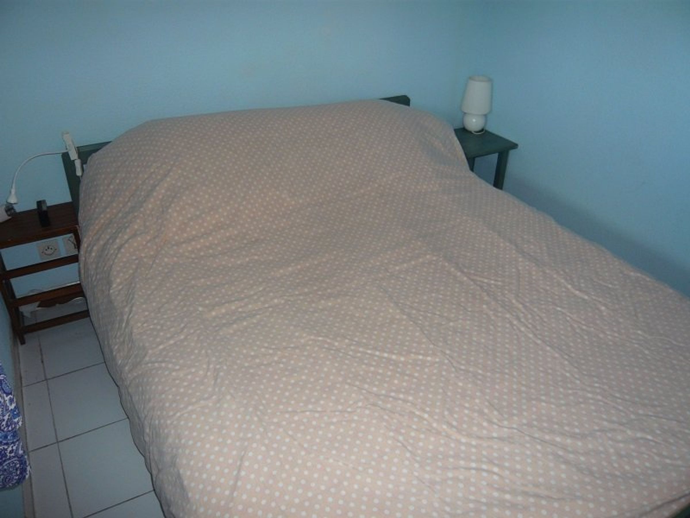 Un lit recouvert d’une housse à pois, une table de chevet avec une lampe et une prise électrique au mur.