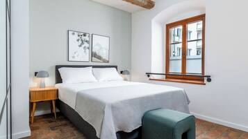 A bedroom with a bed, two bedside tables with lamps, a bench, a window with a view of buildings, and a brick floor.