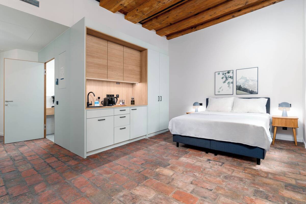 Een slaapkamer met een stenen vloer, een bed met witte beddengoed, een houten plafond, een keuken met witte kasten, en twee ingelijste foto’s aan de muur.