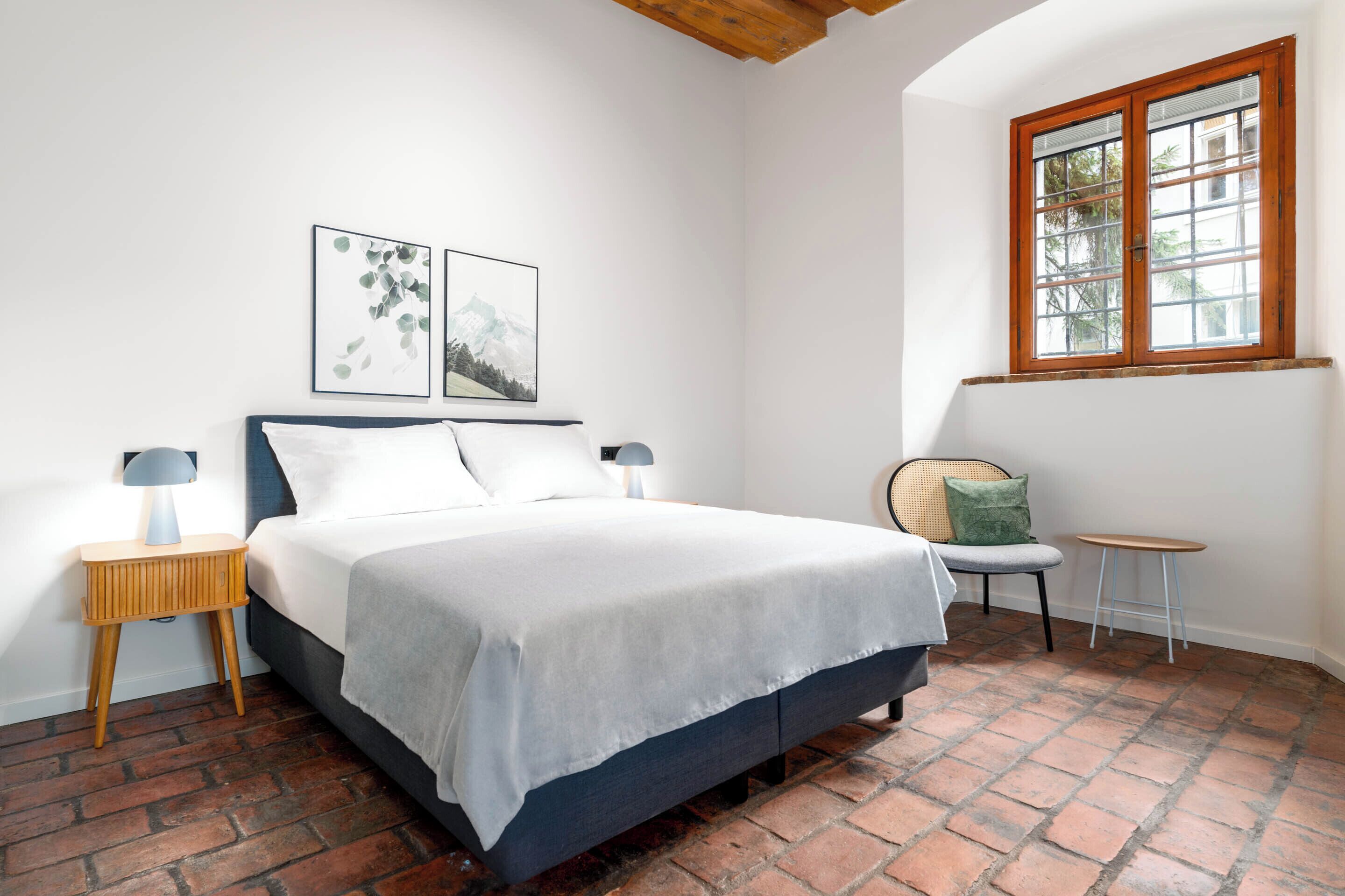 A bedroom with a bed, two bedside tables, a chair, a small table, and a window.