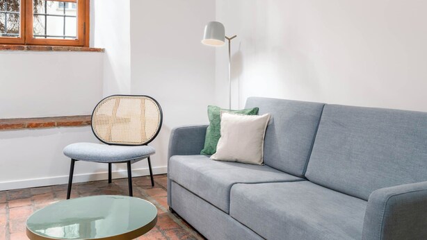 Een kamer met een grijze bank, een ronde glazen tafel, een rieten stoel, een vloerlamp en een raam met houten kozijn.