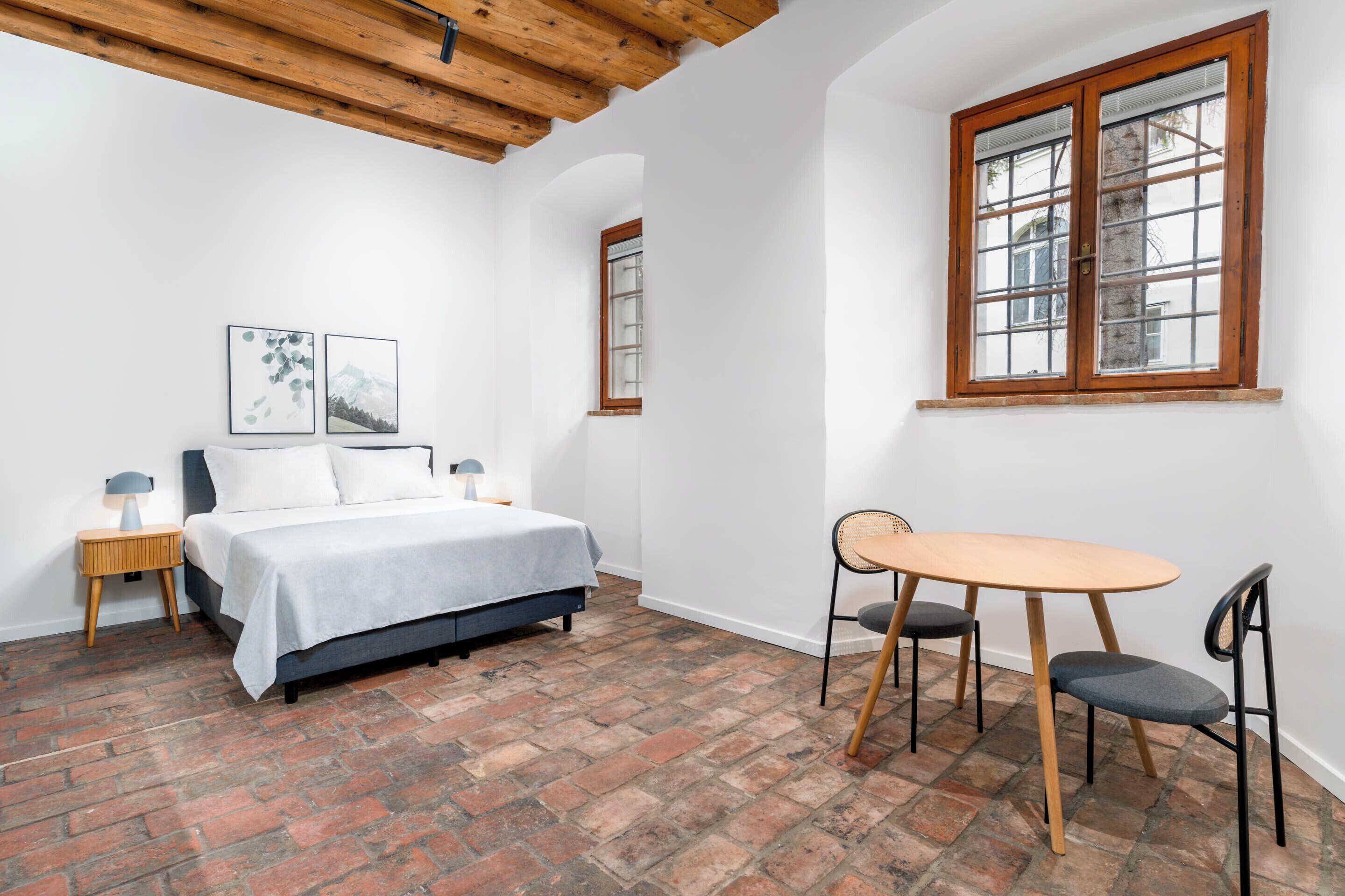 A bedroom with a bed, a small table with two chairs, a window with wooden frames, and a brick floor.
