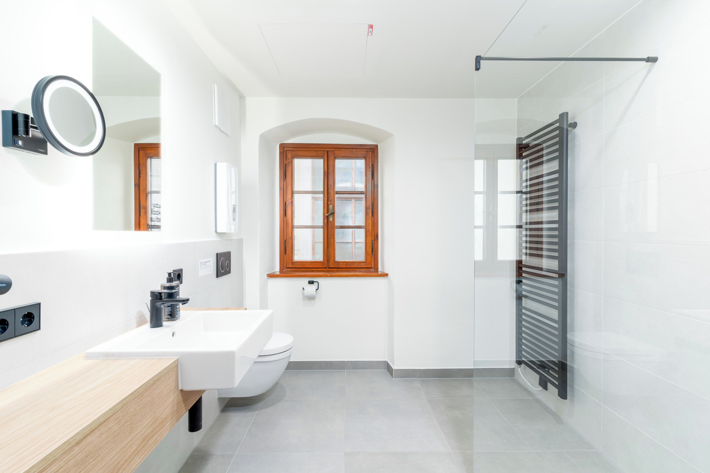 A modern bathroom with a white sink, a mirror, a toilet, and a window with wooden frames.