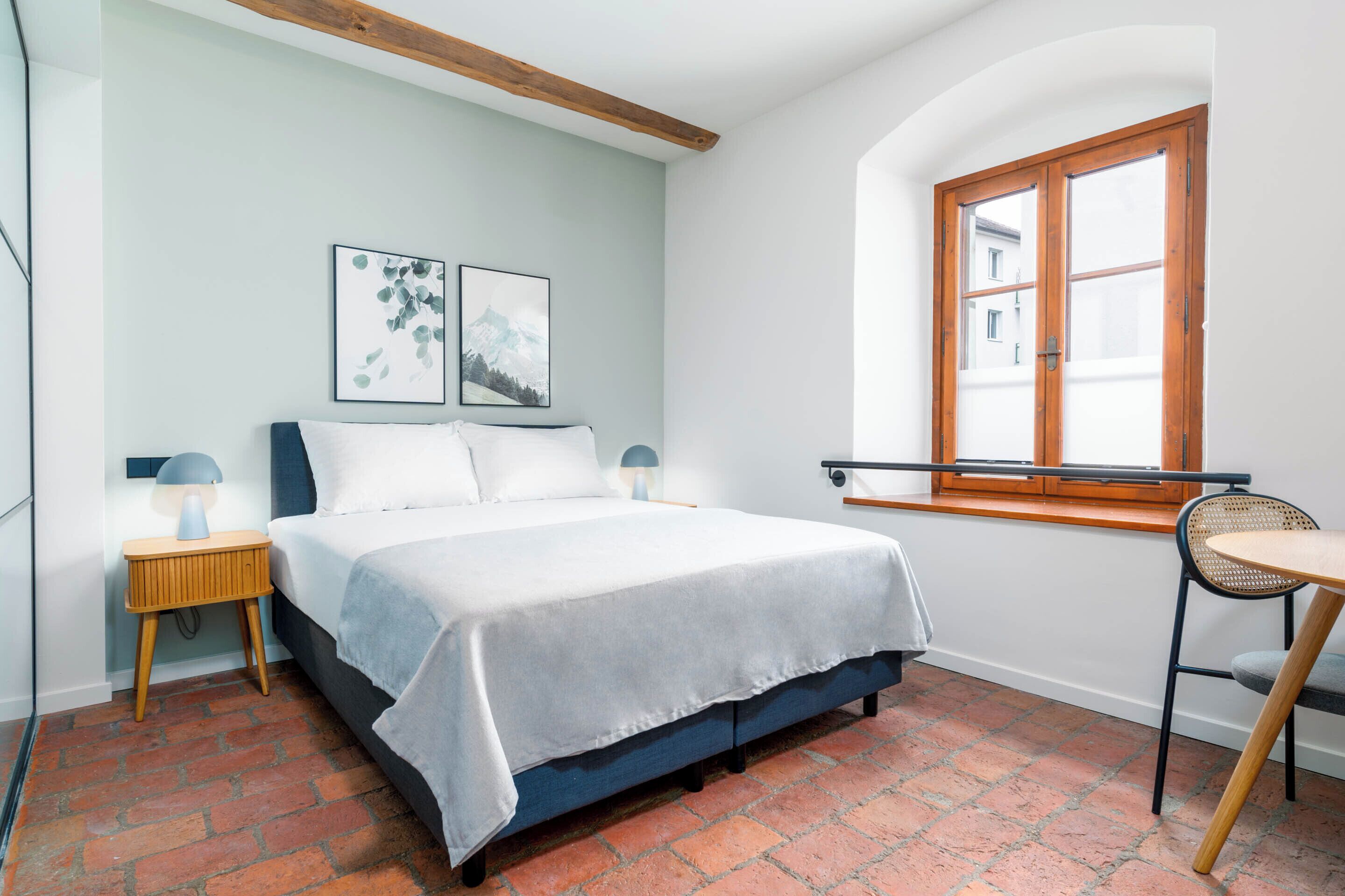 A bedroom with a bed, a nightstand, a chair, a window, and artwork on the wall.