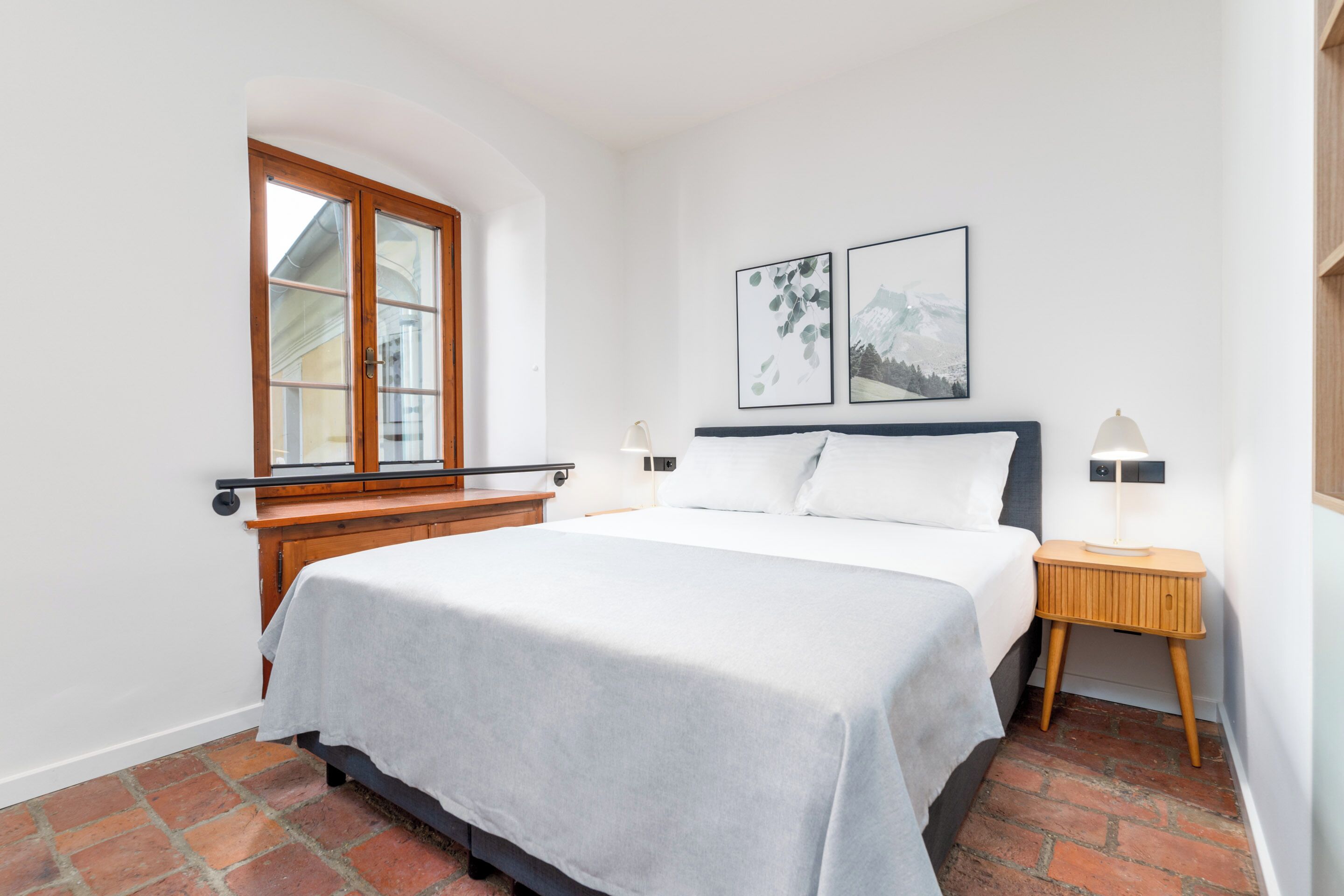Un dormitorio con una cama, una ventana, una mesita de noche y dos cuadros colgados en la pared.