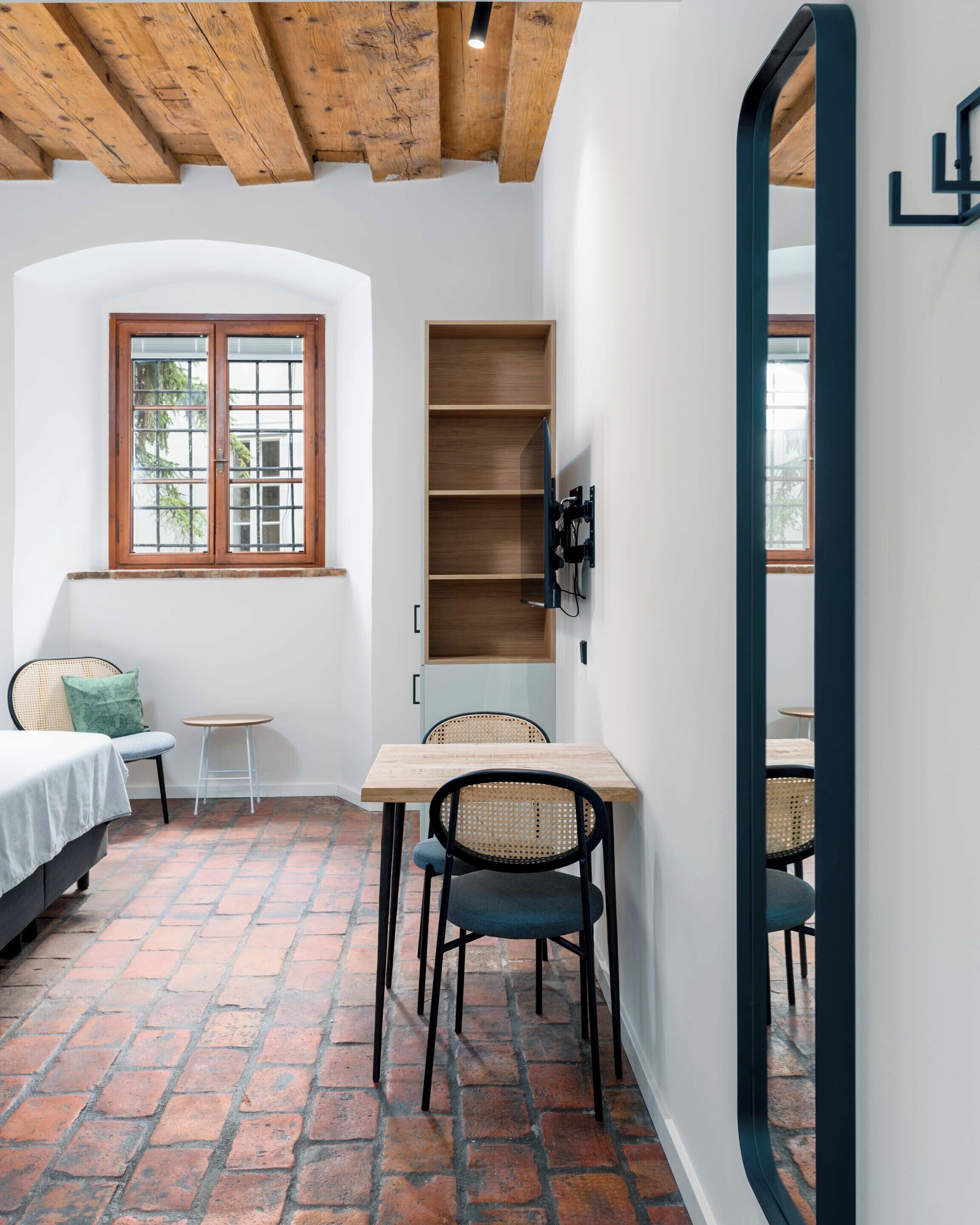 Habitación compacta con suelo de ladrillo, una cama, una mesita con sillas, una estantería de madera y una ventana con vistas a la vegetación.
