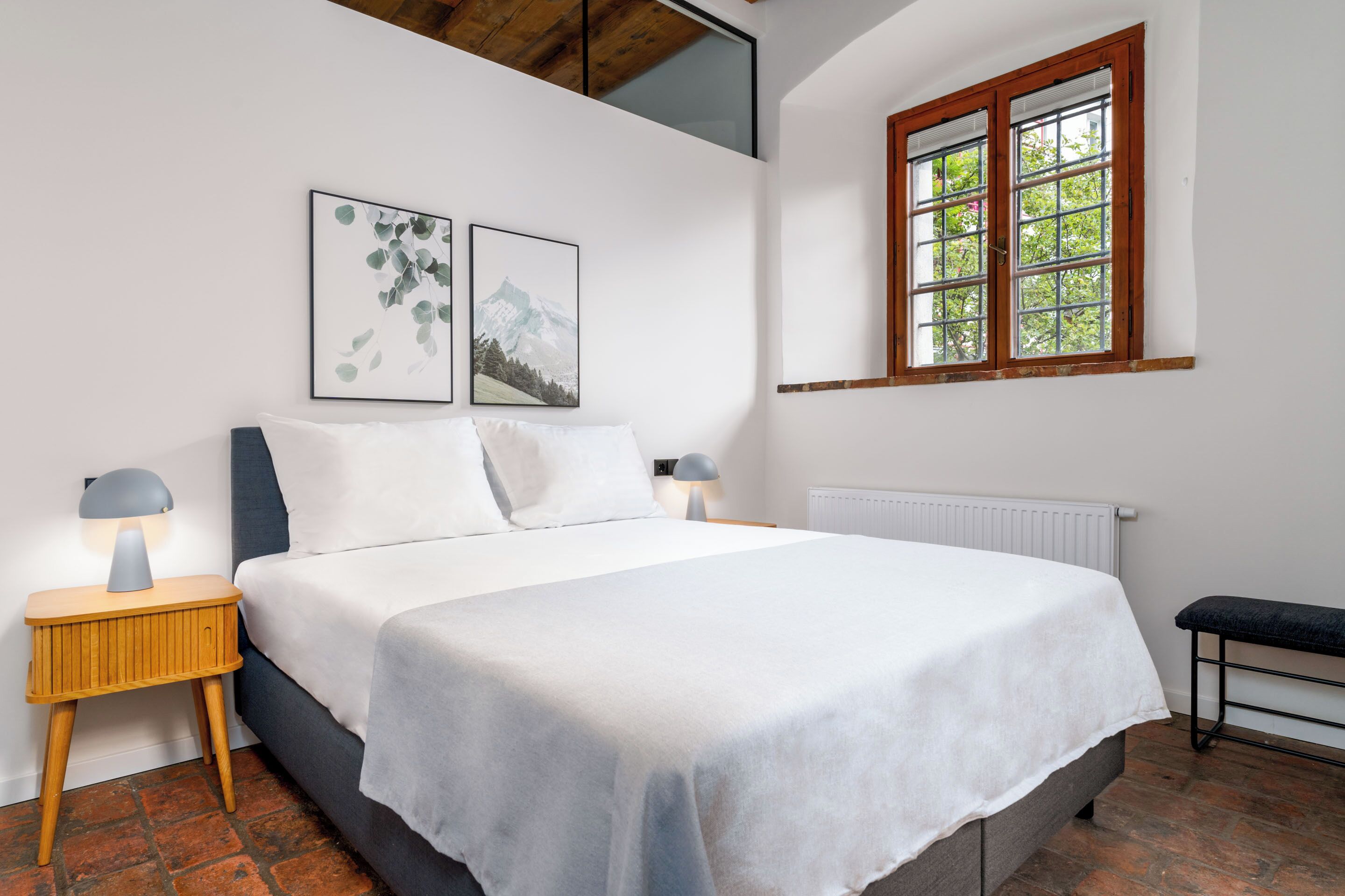 A bedroom with a bed, two wall-mounted pictures, a window, a bedside table with a lamp, and a bench.